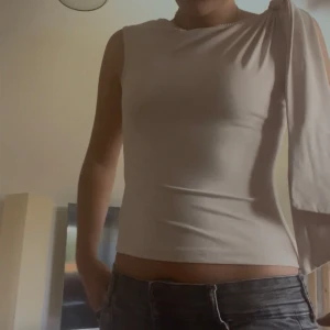 Beige oneshoulder topp med knyt - Säljer en beige, tight oneshoulder topp med cool knytning på ena axeln. Toppen är ärmlös på ena sidan och har en lång tygdel som hänger ner från axeln, tyget är också väldigt stretchigt och bekvämt. Perfekt för sommaren