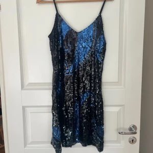 Svart och blå paljettklänning - Säljer en kort svart paljettklänning med blått mönster från märket Manér. Klänningen har smala axelband och v-ringning. Storlek small/medium. Aldrig använd. Sitter snyggt och har en riktigt festlig vibe!