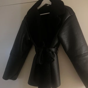Perfekt höstjacka - Snygg svart skinnjacka med bred krage i teddy och knytbälte i midjan. Jackan har en loose passform och långa ärmar, perfekt för kalla dagar. Materialet är en mix av skinn och teddyfoder för extra värme och stil. Passar perfekt nu till hösten/tidig vinter. Liknar den populära jackan från Adoore. Slutsåld på hemsidan. 