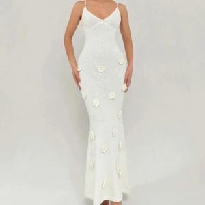 Vit långklänning med blommor och öppen rygg - Supersnygg vit långklänning med smala axelband, öppen rygg och knyt i ryggen. Klänningen har figurnära passform och är dekorerad med 3D-blommor framtill. Perfekt för sommarens alla tillfällen!