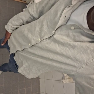 Ljusgrå linneskjorta med knappar - Säljer en stilren ljusgrå skjorta i linne med klassisk krage och vita knappar. Skjortan har lång ärm och en avslappnad passform, perfekt att bära öppen över en t-shirt för en chill look. Helt nytt