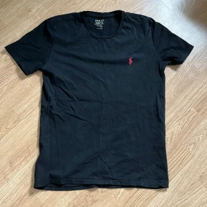 Svart t-shirt från Polo Ralph Lauren - Svart t-shirt från Polo Ralph Lauren i 100% bomull. Klassisk passform med korta ärmar och rund hals. På bröstet finns den ikoniska röda broderade polospelaren. Enkel och stilren design som funkar till allt.