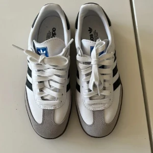 Adidas Samba vita  - Säljer ett par klassiska Adidas Samba sneakers i vitt med svarta ränder och grå mockadetaljer vid tån. Skorna är använda en gång och är i ett mycket fint skick! Ord pris 1 449kr.