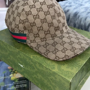 Gucci keps med GG-mönster och röd/grön detalj - Exklusiv keps från Gucci i beige och brunt med klassiskt GG-monogram över hela kepsen. Sidorna har den ikoniska röd-gröna randen och justerbar rem bak i brunt skinn. Perfekt accessoar för dig som vill sticka ut med lyxig streetstyle.