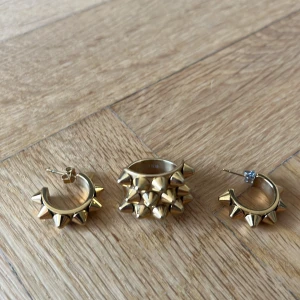 Guldiga smycken med nitar - Säljer ett set med chunky guldfärgade smycken: ett par örhängen och en ring, alla dekorerade med coola konformade nitar. Ger en edgy vibe och sticker verkligen ut. Perfekt för dig som gillar statement-accessoarer.