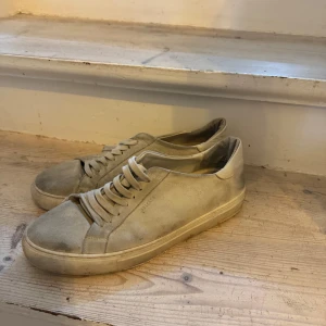 Beige sneakers från ARIGATO i mocka - Snygga beige sneakers från ARIGATO i mjuk mocka med klassisk låg profil och rund tå. Skorna har platt sula och snörning framtill, perfekt för en clean och stilren look. Diskret logga på sidan och minimalistisk design.