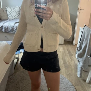 Beige stickad kofta med fickor från Gina Tricot  - Superfin beige stickad kofta med rund halsringning och snygga guldknappar framtill. Två praktiska fickor framtill och ribbade muddar vid ärmslut och nederkant. Perfekt att slänga över en topp för en chill look. Koftan saknar en knapp