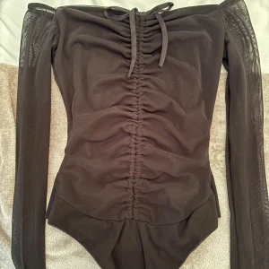  Body från PrettyLittleThing - Säljer en svart bodysuit i mesh från PrettyLittleThing med långa meshärmar och snygg rynkdetalj framtill. Offshoulder-modell med knyt upptill och tryckknappar i grenen. Perfekt för dig som vill ha en trendig och figurnära topp med det lilla extra. Passar mer som S.
