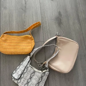 Tre trendiga axelväskor i olika färger - Tre snygga axelväskor: en gul med croco-mönster, en beige i glansigt material och en vit med ormmönster och silverdetaljer. Perfekta för att lyfta din outfit och rymmer det viktigaste du behöver ha med dig.