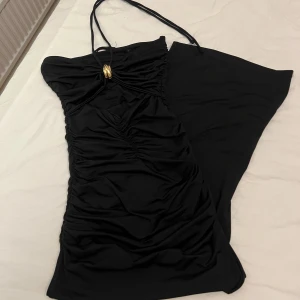 Svart långklänning från Zara med cut out - Supersnygg svart långklänning från Zara med halterneck och gulddetalj framtill. Klänningen har cut out vid bysten, rynkade detaljer och är figurnära. Perfekt för dig som vill sticka ut med en elegant och trendig look.