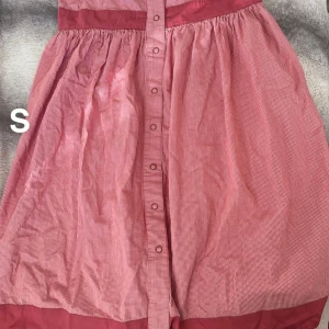 Rutig röd kortklänning med knappar - Supersöt röd och vit rutig kortklänning med knappar hela vägen framtill och markerad midja. Klänningen har en lätt utställd kjol och bredare röd kant upptill och nertill. Perfekt för dig som gillar retrovibbar och vill sticka ut lite extra.🌸Fint använt skick. Svårt att beskriva färgen men typ hallonrosa. Storlek s