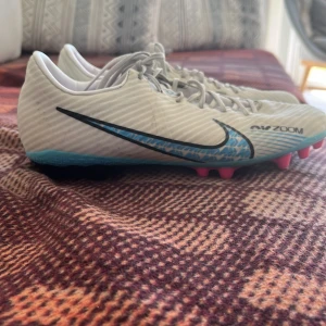 Nike Air Zoom fotbollsskor vita/blå - Säljer ett par Nike Air Zoom fotbollsskor i vitt med blå och svarta detaljer. Skorna har snörning, rosa innersula och mönstrad blå sula med dobbar. Fotbollsskorna är hela och rena och endast använda några månader.