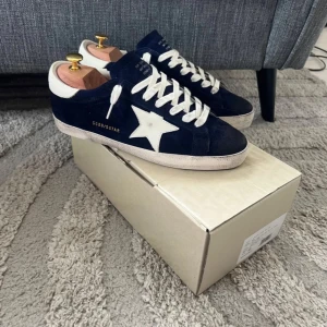 Golden Goose Superstar sneakers blå/vit - Golden Goose Superstar sneakers i mörkblå mocka med vit stjärndetalj på sidan, vita skosnören och vit sula. Klassisk låg modell med rund tå och retrovibbar. Märkeslogga i guld på sidan och häl. Kommer med originalkartong och dustbag.