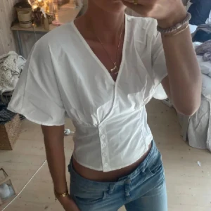 Vit omlott blus med knappar - Säljer en vit blus med v-ringning och omlottkänsla. Toppen har korta, vida ärmar och knappar framtill som ger en snygg, figurnära passform. Perfekt till jeans för en clean och fräsch look. Vet inte vart den är ifrån tyvärr då lappen är bortklippt 