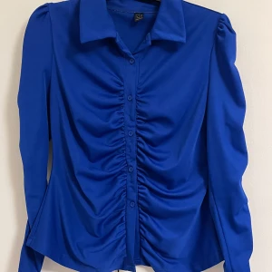 Kornblå skjorta med rynkade detaljer - Snygg kornblå skjorta med långa ärmar och rynkade detaljer längs knappraden framtill. Skjortan har klassisk krage och en figurnära passform som ger en trendig look. Clasy och sexig samtidigt. Bra skick! 