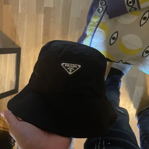 Svart bucket hat från Prada - Snygg svart bucket hat från Prada med ikonisk metallogo framtill. Klassisk form med bredare brätte och tillverkad i glansig syntet. Perfekt accessoar för att lyfta din outfit med en lyxig touch.