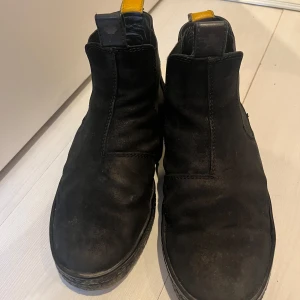 Svarta chelsea boots från Canada Snow - Svarta chelsea boots i mocka från Canada Snow med rund tå och platt sula. Snygga detaljer med svarta resårpaneler på sidorna och gul dragflik bak. Perfekta för dig som gillar stilrena och klassiska boots med en twist.