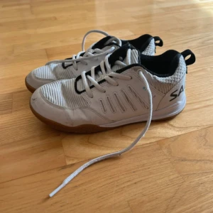  Salming rival 2 storlek 35 - Vita inomhusskor från Salming med svarta och grå detaljer. Skorna har snörning, mönstrad ovandel och gummisula för bra grepp. Perfekta för träning eller sport inomhus. Bekväm passform och sportig look. Har en liten defekt men inget som märks av när man använder dem 💞