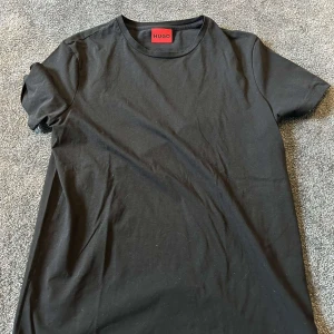 Svart t-shirt från Hugo Boss - En clean svart t-shirt från Hugo Boss med klassisk rund hals och korta ärmar. T-shirten är i mjuk bomull och har en enkel, stilren look utan tryck eller mönster. Perfekt för dig som gillar minimalistisk stil och vill ha ett snyggt basplagg i garderoben.