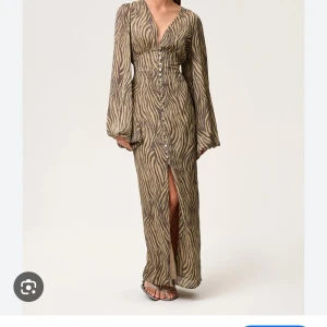 Adorre  - Snygg långklänning med zebramönster i beige och brunt. Djup v-ringning, långa vida ärmar och knappar hela vägen framtill. Klänningen har en slits framtill och markerad midja för en trendig siluett. 