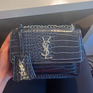 Marinblå YSL axelväska i skinn - Säljer en mörkblå axelväska från YSL i croco-präglat skinn med silverfärgad kedja och ikonisk YSL-logga framtill. Väskan har en stilren, boxig form. Remmen kan användas som både kort och liten. Det är en k*pia💞
