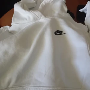 Vit hoodie från Nike - Säljer en klassisk vit hoodie från Nike med svart logga på bröstet. Tröjan har huva och är perfekt för en avslappnad stil. Passar både till jeans och träningsbyxor.