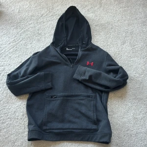 Mörkgrå hoodie från Under Armour - Säljer en mörkgrå hoodie från Under Armour med dragkedja vid halsen och röd logga på bröstet. Tröjan har huva med dragsko och en stor ficka framtill med dragkedja. Perfekt för träning eller chill.