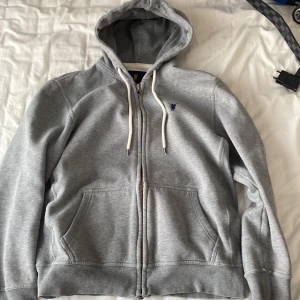 Grå hoodie med dragkedja från polo - Säljer en klassisk grå hoodie från Ralph Lauren med dragkedja och huva. Tröjan har två fickor framtill, vita snören i huvan och en liten broderad logga på bröstet. Perfekt för en avslappnad stil.