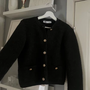 Svart cardigan jacka från Zara - Stilren svart cardigan jacka från Zara med guldknappar framtill och på fickorna. Jackan har rund halsringning och två fickor framtill. Perfekt för att lyfta en enkel outfit.