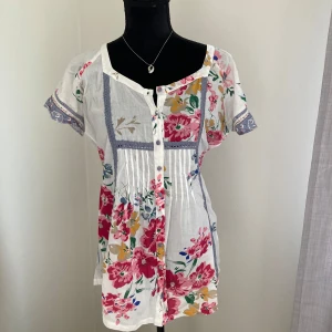 Vit blommig blus med knappar och knytband - Säljer en söt vit blus med färgglatt blommönster i rosa, blått och gult. Blusen har korta ärmar med spetsdetaljer, knappar framtill och knytband i midjan. Perfekt för vår och sommar!