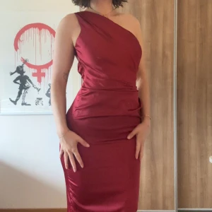 Vinröd one shoulder långklänning från Babyboo - Säljer en vinröd långklänning i satin från Babyboo. Klänningen har en elegant one shoulder-design och öppen rygg. Snygg figurnära passform som framhäver siluetten. Perfekt för speciella tillfällen.