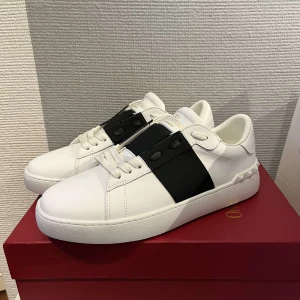 Valentino Garavani vita sneakers med svart detalj - Säljer ett par vita Valentino Garavani sneakers med bred svart rem över mitten och ikoniska nitar på hälen. Skorna har snörning och är tillverkade i skinn. Perfekta för dig som vill ha en stilren och lyxig look.