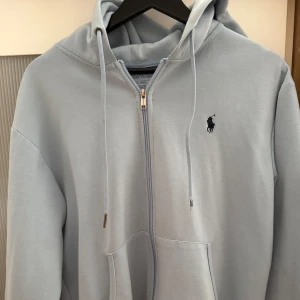 Ralph Lauren zip - Säljer en ljusblå Polo Ralph Lauren-huvtröja, perfekt för sommaren! Denna klassiska huvtröja är tillverkad av mjukt och lätt material, idealiskt för svala sommarkvällar eller kyliga morgnar. Den ljusblå färgen ger en fräsch och somrig känsla, och den ikoniska Polo-logotypen ger en touch av stil.