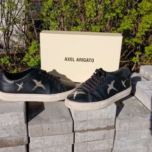 Svarta sneakers med fågelmotiv från Axel Arigato - Snygga svarta sneakers från Axel Arigato med vita fågelmotiv på sidorna. Skorna har svarta snören och vit platt sula. Perfekta för dig som gillar stilrena men unika detaljer. Kommer med originalkartong.