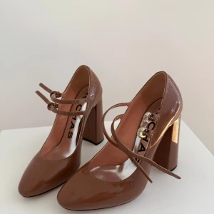 Brun/rosa lackade klackskor från Rochas - Säljer ett par brun/rosa lackade klackskor från Rochas med rund tå och dubbla remmar över foten. Skorna har hög blockklack och en elegant, glansig finish. Perfekta för dig som vill sticka ut med stilrena detaljer och lyxig känsla. Endast använda en gång. 