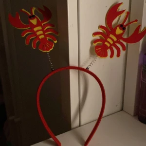 Rött kräftdiadem med fjädrande dekorationer - Roligt diadem med två röda kräftor på fjädrar som sticker upp. Perfekt accessoar till kräftskivan eller temafesten. Diademet är täckt i rött tyg och kräftorna är gjorda i filtliknande material.