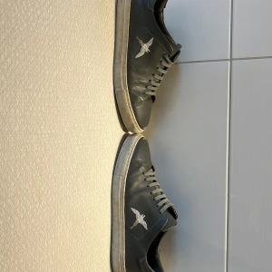 Svarta sneakers med fågelbrodyr - Säljer ett par svarta sneakers med grå snörning och en broderad vit fågel på sidan. Skorna har platt sula och rund tå, perfekt för en stilren vardagslook. I dåligt skick men fungerar bra ändå