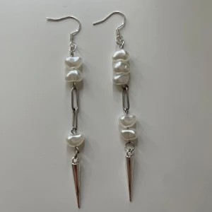 Långa örhängen med pärlor och silverfärgade spikes - Unika örhängen med vita pärlor i hjärtform och avlånga silverfärgade spikes. Örhängena har en modern design med metallkedja och passar perfekt för dig som vill sticka ut.