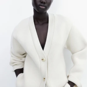 Vit stickad kofta med knappar - Säljer en stilren vit stickad kofta med djup v-ringning och tre knappar framtill. Koftan har långa ärmar och en oversized passform, perfekt för lager-på-lager. Enkel och tidlös design som passar till mycket.