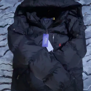 Svart pufferjacka från Polo Ralph Lauren - Säljer en svart pufferjacka från Polo Ralph Lauren med röd broderad logga på bröstet. Jackan har dragkedja framtill och hög krage. Perfekt för kyliga dagar och riktigt snygg till streetwear-stilen. S den passar M 