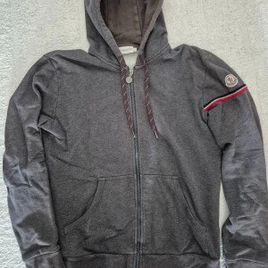 Moncler Maglia Cardigan Zip Hoodie L - Säljer en mörkgrå hoodie från Moncler med dragkedja framtill och huva. Tröjan har fickor på magen, snören i huvan och Moncler-logga på ärmen tillsammans med en röd och vit rand. Perfekt för en avslappnad stil. Små i storleken, mer som en M.