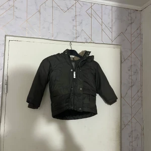 Mörkgrön vinterjacka med foder från Lindex - Mysig mörkgrön vinterjacka från Lindex med mjukt teddyfoder i huvan. Jackan har dragkedja och knappar framtill samt två fickor. Perfekt för kalla dagar och håller värmen bra.