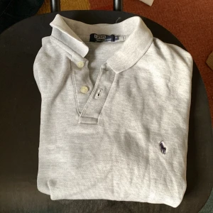 Ljusgrå pikétröja från Ralph Lauren - Säljer en klassisk ljusgrå pikétröja från Ralph Lauren med den ikoniska broderade loggan på bröstet. Tröjan har krage, knappar framtill och är tillverkad i bomull. Perfekt för en stilren och avslappnad look.