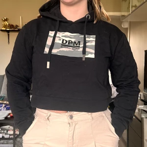 Svart croppad hoodie från Björn Borg - Svart croppad hoodie från Björn Borg med kamouflagemönstrad logga på bröstet och justerbar huva med snören. Perfekt för en avslappnad och cool stil.