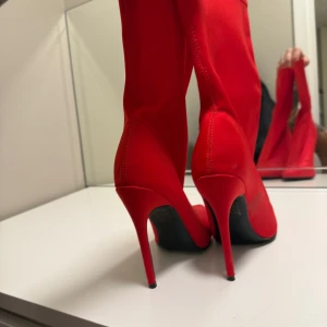 Röda stretchiga boots med klack - Säljer ett par knallröda boots med hög smal klack och spetsig tå. Skorna är i ett stretchigt syntetmaterial som sitter tight runt benet och har en modern, stilren look. Perfekta för dig som vill sticka ut med färg.