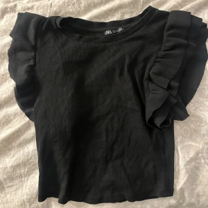 Svart volangtopp från Zara - Svart topp från Zara med volangdetaljer på axlarna. Modellen är croppad och har korta ärmar. Perfekt för en trendig och stilren look. (Storlek M men passar mig i Xs också)  
