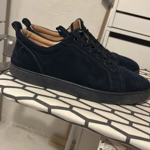 louboutin skor navy blue - Snygga svarta sneakers i mocka med svarta snören och låg profil. Skorna har en rund tå och platt sula, samt en röd undersida på sulan som ger en cool kontrast. Insidan är ljusbeige och de har diskreta detaljer på sidan.