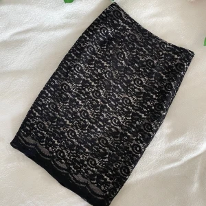 Svart spetskjol från Oxygène - Säljer en svart, knälång kjol från Oxygène med vacker blommig spets över ett ljust innerfoder. Kjolen har en klassisk, rak passform och är perfekt för dig som gillar stilrena detaljer och elegant look. Längd: 59cm, Midja: 34cm