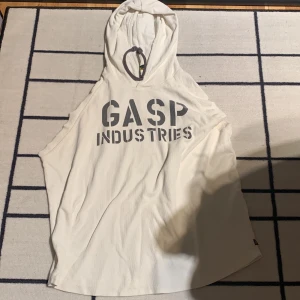Vit ärmlös hoodie från GASP Industries - Säljer en vit ärmlös hoodie från GASP Industries i storlek L. Tröjan har tryck både fram och bak, samt en huva med snörning. Perfekt för gymmet eller träning. Materialet känns mjukt och bekvämt.