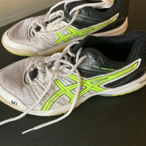 Vita och svarta Asics Gel-Rocket träningsskor - Säljer ett par Asics Gel-Rocket träningsskor i vitt och svart med neongula detaljer. Skorna har snörning och en ventilerande mesh-överdel. Perfekta för inomhusträning eller sport.